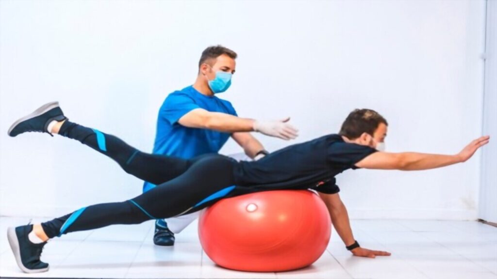 Physiotherapist with Mask Helps Patient Stretching On Ball
